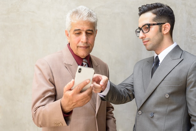 Persiano padre e figlio come uomini d'affari utilizzando il telefono insieme contro il muro di cemento all'aperto