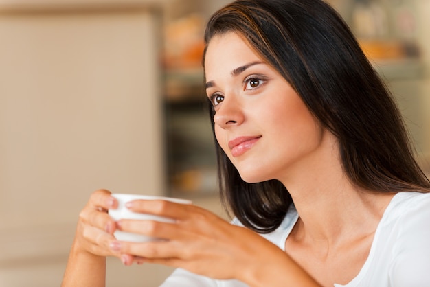 Perso nei pensieri. Giovane donna premurosa che tiene la tazza di caffè e distoglie lo sguardo mentre è seduta nella caffetteria