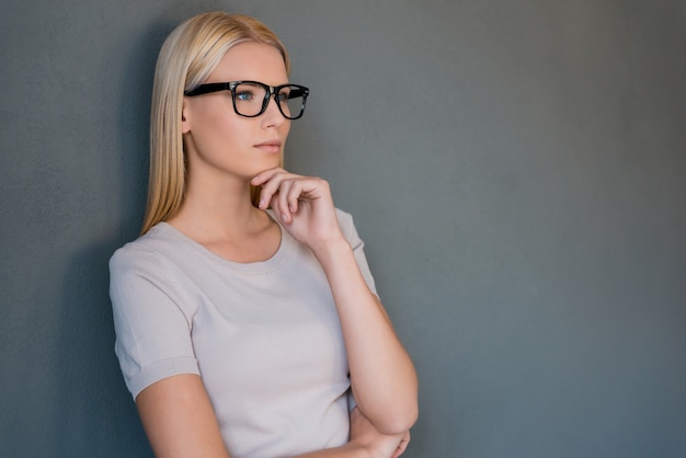 Perso nei pensieri. Giovane donna premurosa dei capelli biondi che tiene la mano sul mento e guarda lontano mentre sta in piedi su uno sfondo grigio