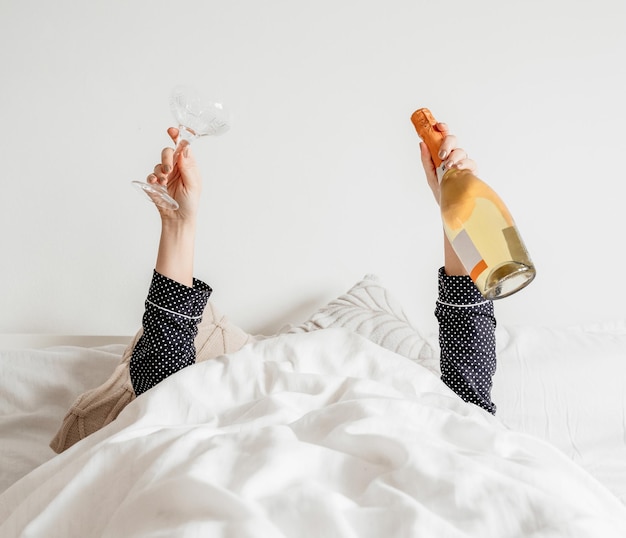 Persona a letto, coperta di lino bianco, con in mano una bottiglia di champagne e un bicchiere.