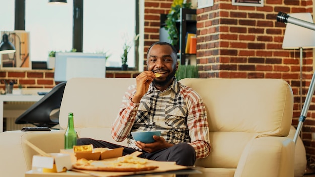 Persona afroamericana che apre la televisione per trovare film, mangiare patatine in una ciotola e bere bottiglie di birra. Giovane ragazzo allegro che si abbuffa guardando il programma televisivo preferito, consumando pasti diversi. Scatto da treppiede.