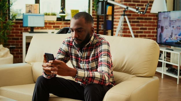 Persona annoiata che scorre senza pensare sul telefono ascoltando il rumore di sottofondo della TV uomo afroamericano che passa il tempo sui social media mentre è stanco nel salotto di un appartamento accogliente e rilassante