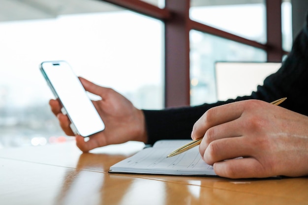 Persona che tiene in mano uno smartphone e scrive in una cartella con una penna d'oro su un tavolo di legno vicino a una finestra Moderno spazio di lavoro multitasking e concetto di pianificazione digitale in un ambiente professionale