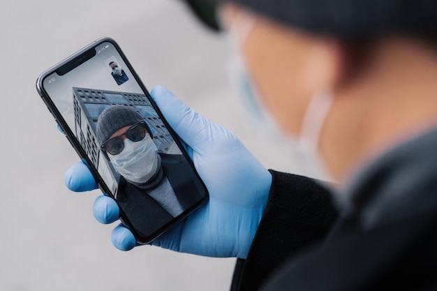 Persona con guanti e maschera che tiene un telefono durante una videochiamata che riflette la nuova normalità della comunicazione nei tempi della pandemia