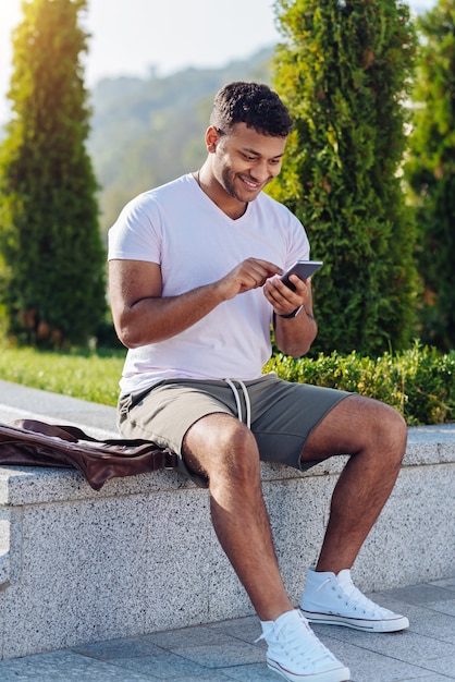 Persona di sesso maschile felice positiva seduta fuori e chinando la testa mentre gioca con il telefono cellulare