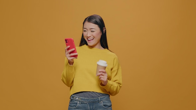 Persona felice che tiene smartphone e tazza di caffè, sorridente in studio. Donna allegra che guarda lo schermo del telefono cellulare e ha una bevanda calda in mano davanti alla fotocamera. Navigazione in internet