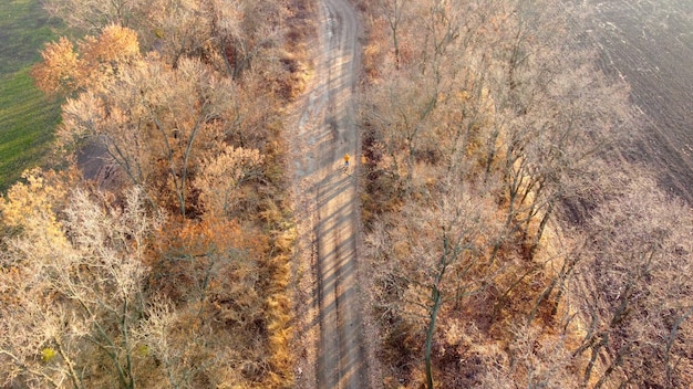 Persona in giacca arancione in piedi con la bicicletta su strada di campagna sterrata tra alberi senza foglie su a