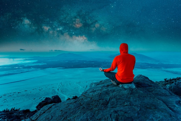 Persona sconosciuta seduta sulla cima della montagna che medita di notte con lo sfondo della Via Lattea