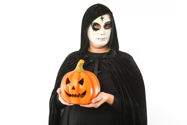 Persona vestita di maschera bianca con croce sulla fronte e mantello di velluto nero con cappuccio, che tiene una zucca jack-o-lantern, su sfondo bianco. Concetto di Carnevale, Halloween e Giorno dei Morti.