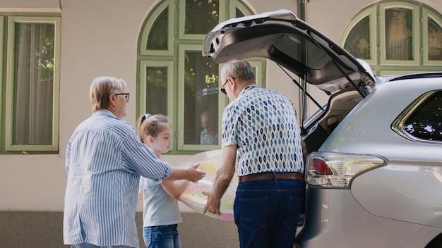 Persone anziane con la nipote che mettono bagagli e carrelli da viaggio all'interno del veicolo mentre vanno in gita estiva. I nonni portano la nipote in spiaggia mentre caricano i bagagli nel bagagliaio dell'auto