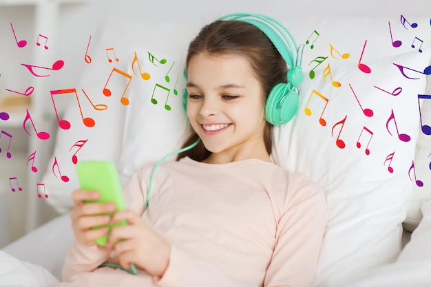 persone, bambini e concetto di tecnologia - felice ragazza sorridente sdraiata sveglia con smartphone e cuffie a letto ascoltando musica a casa su note musicali