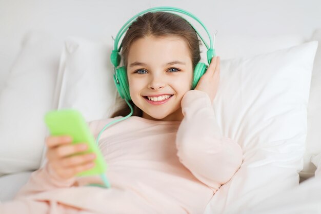 persone, bambini, riposo e concetto di tecnologia - felice ragazza sorridente sdraiata sveglia con smartphone e cuffie a letto ascoltando musica a casa