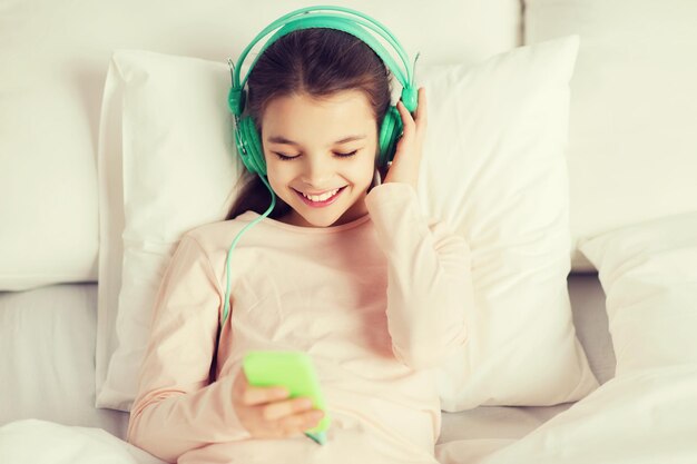 persone, bambini, riposo e concetto di tecnologia - felice ragazza sorridente sdraiata sveglia con smartphone e cuffie a letto ascoltando musica a casa