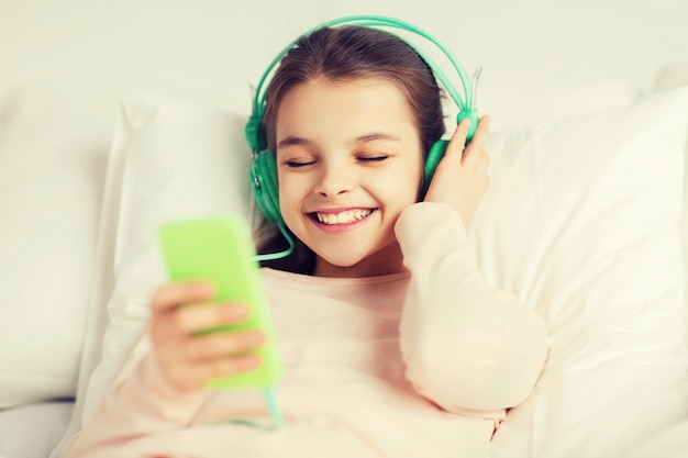 persone, bambini, riposo e concetto di tecnologia - felice ragazza sorridente sdraiata sveglia con smartphone e cuffie a letto ascoltando musica a casa