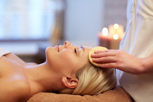 persone, bellezza, spa, stile di vita sano e concetto di relax - primo piano di bella giovane donna sdraiata con gli occhi chiusi e con massaggio viso con spugna nella spa