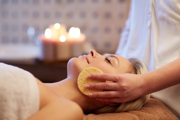 persone, bellezza, spa, stile di vita sano e concetto di relax - primo piano di bella giovane donna sdraiata con gli occhi chiusi e con massaggio viso con spugna nella spa