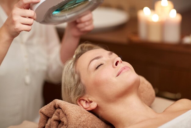 persone, bellezza, spa, stile di vita sano e concetto di relax - primo piano di bella giovane donna sdraiata con gli occhi chiusi e cosmetologo che guarda attraverso la lampada d'ingrandimento nella spa