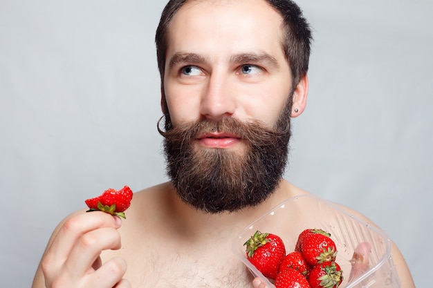 Persone, cibo, bellezza, stile di vita, concetto di San Valentino - ritratto del primo piano giovane che tiene una fragola e sorridente. Ritratto di bello barbuto e baffi. su sfondo grigio