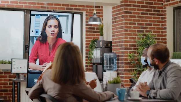 Persone con maschera facciale che parlano in teleconferenza con una donna, utilizzando la videoconferenza online sul monitor. Colleghi che fanno telecomunicazioni in videoconferenza remota durante la pandemia.