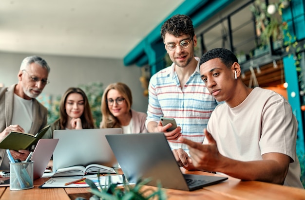 Persone creative multirazziali in un ufficio moderno. Un gruppo di giovani uomini d'affari e capo senior stanno lavorando insieme a laptop, tablet, smartphone, notebook, grafici. Team di successo nel coworking