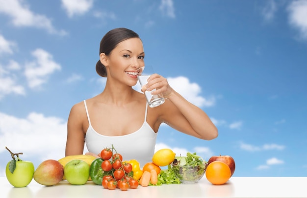 persone, dieta, alimentazione sana e concetto di cibo - donna felice che beve acqua da vetro, frutta e verdura su sfondo blu cielo e nuvole