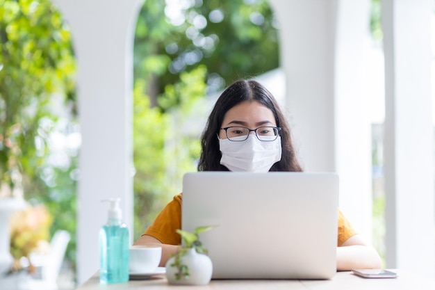Persone freelance donna d'affari che indossa una maschera protettiva che lavora casualmente con un computer portatile in una caffetteria come lo sfondo, che lavora da casa, concetto di prevenzione della diffusione del virus COVID-19