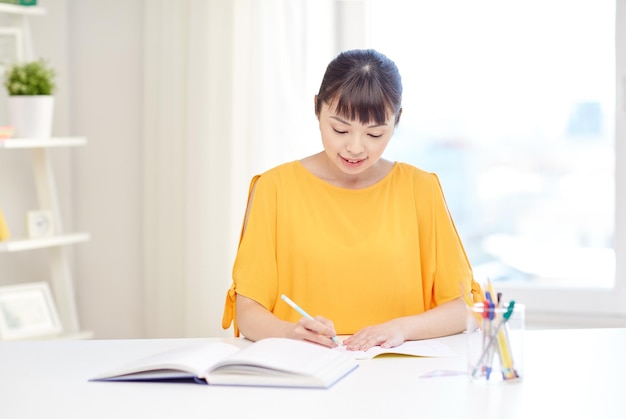 persone, istruzione, scuola superiore e concetto di apprendimento - felice giovane studentessa asiatica con libro e blocco note che scrive a casa