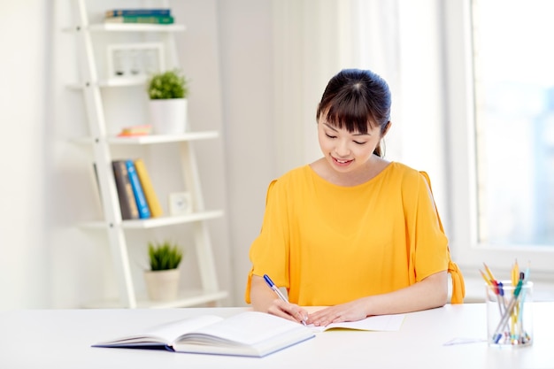 persone, istruzione, scuola superiore e concetto di apprendimento - felice giovane studentessa asiatica con libro e blocco note che scrive a casa