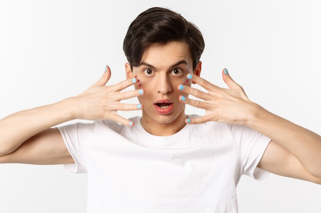 Persone, lgbtq e concetto di bellezza. Primo piano di un bellissimo uomo gay con glitter sul viso, che mostra lo smalto sulle unghie, guardando impertinente alla telecamera, in piedi su sfondo bianco.