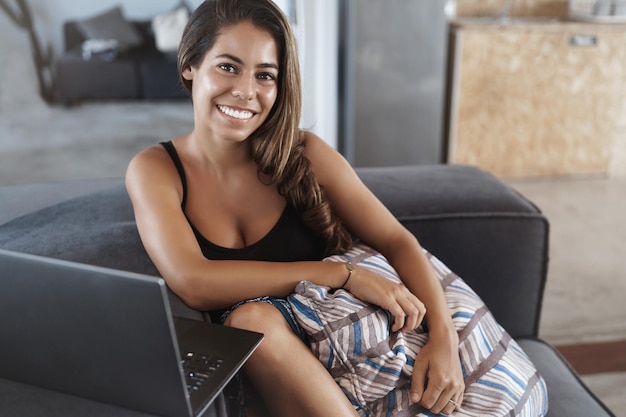 Persone nomadi digitali e concetto di business Attraente giovane donna allegra seduta sul divano che abbraccia il cuscino e sorridente fotocamera guardando video laptop che lavora a distanza dallo spazio di coworking
