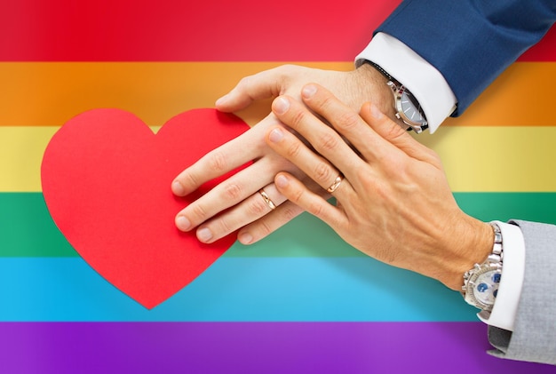 persone, omosessualità, matrimonio omosessuale, San Valentino e concetto di amore - primo piano di mani di coppia gay maschio sposata felice con forma di cuore di carta rossa su sfondo bandiera arcobaleno