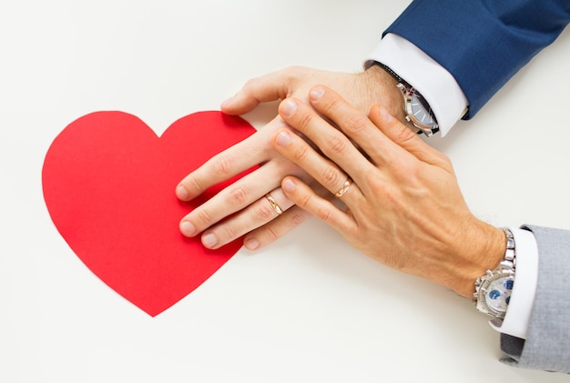 persone, omosessualità, matrimonio omosessuale, San Valentino e concetto di amore - primo piano di mani di coppia gay maschio sposata felice con forma di cuore di carta rossa sul tavolo