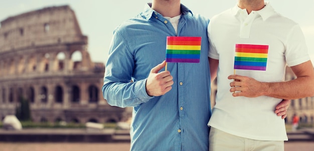 persone, omosessualità, matrimonio tra persone dello stesso sesso, concetto di viaggio e amore - primo piano di coppia gay maschio felice che tiene bandiere arcobaleno e abbraccia dal retro sul Colosseo sullo sfondo di roma