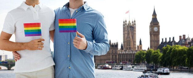 persone, omosessualità, matrimonio tra persone dello stesso sesso, concetto di viaggio e amore - primo piano di una coppia gay maschile che tiene bandiere arcobaleno e si abbraccia da dietro sul big ben e sulla casa del parlamento sullo sfondo di londra