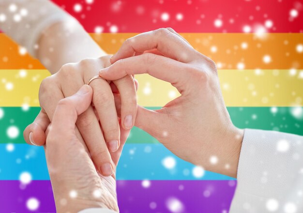 persone, omosessualità, matrimonio tra persone dello stesso sesso e concetto di amore - primo piano delle mani di una coppia lesbica felice che indossano l'anello nuziale su sfondo bandiera arcobaleno sopra l'effetto neve