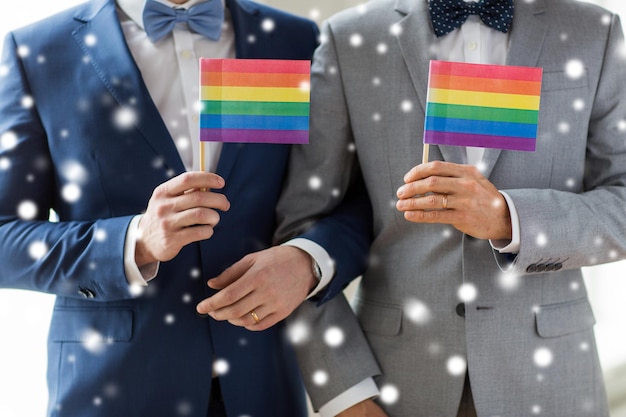 persone, omosessualità, matrimonio tra persone dello stesso sesso e concetto di amore - primo piano di felice coppia gay maschile in giacca e cravatta con fedi nuziali che tengono bandiere arcobaleno sopra l'effetto neve