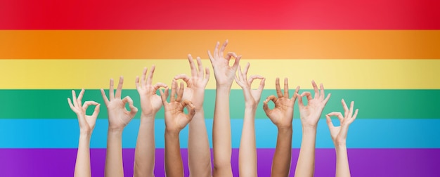 persone, orgoglio gay, gesto e concetto omosessuale - mani umane che mostrano segno ok su sfondo a strisce bandiera arcobaleno