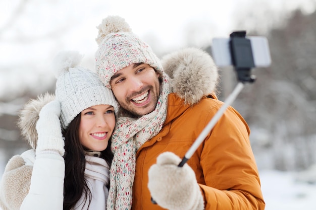 Persone, stagione, amore, tecnologia e concetto di tempo libero - coppia felice che scatta foto con il bastone selfie dello smartphone su sfondo invernale