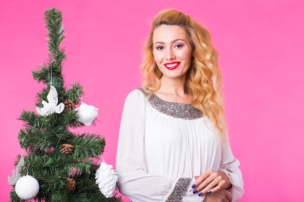Persone, vacanze e concetto di natale - giovane donna bionda in piedi vicino all'albero di Natale sulla parete rosa.