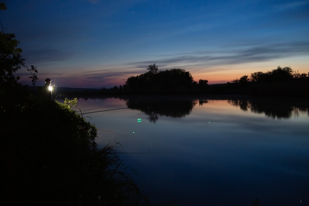 Pesca notturna, pescatore con una canna da pesca in piedi sulla riva del lago.