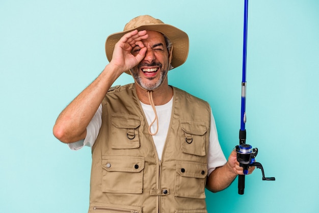 Pescatore caucasico di mezza età che tiene l'asta isolata su sfondo blu eccitato mantenendo il gesto ok sull'occhio.