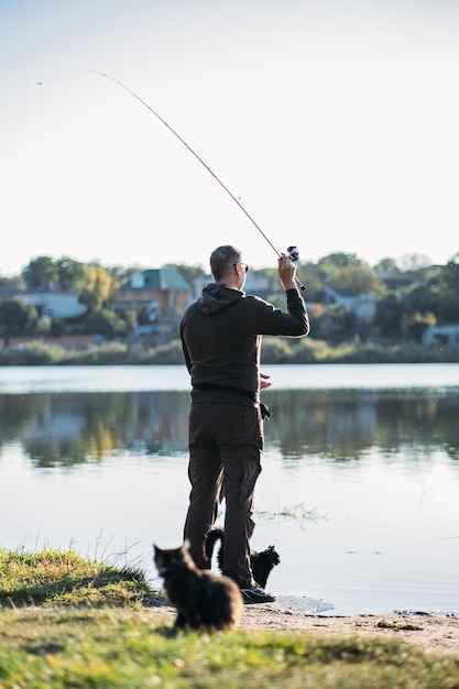Pescatore con canna da spinning e gatto vicino a lui su sfondo naturale pescatore uomo con pesca spinning