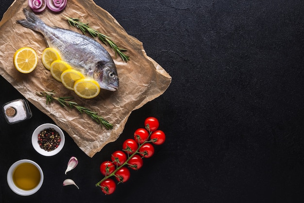 Pesce crudo di Dorado preparato per la cottura con fette di limone, rosmarino, cipolla, pomodorini, spezie e olio su sfondo nero. Vista dall'alto. Copia spazio.