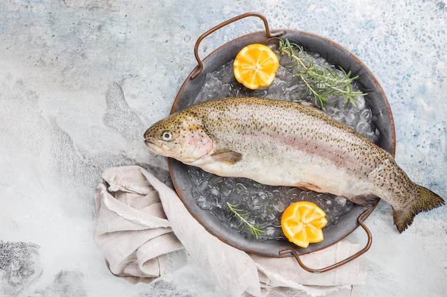 Pesce crudo di trota sul vassoio con ghiaccio con rosmarino e limone su sfondo chiaro di pietra, vista dall'alto