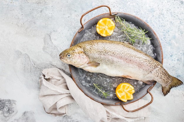 Pesce crudo di trota sul vassoio con ghiaccio con rosmarino e limone su sfondo chiaro di pietra, vista dall'alto