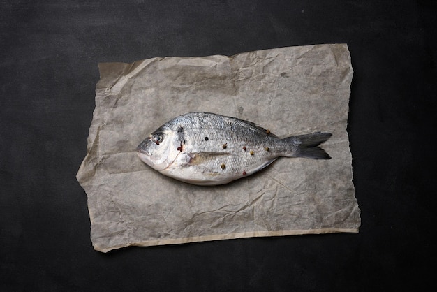 Pesce dorado intero crudo su carta pergamena marrone e spezie per cucinare vista dall'alto sul tavolo nero