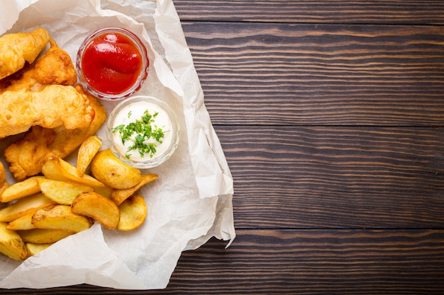 Pesce e patatine fritte tradizionali britanniche degli alimenti a rapida preparazione con i tuffi assortiti per scelta, su fondo di legno rustico della carta