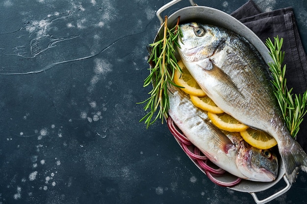 Pesce fresco crudo dorado o orata con ingredienti per fare limone, timo, aglio, pomodorini e sale su uno sfondo di ardesia nera, pietra o cemento. Vista dall'alto con copia spazio.