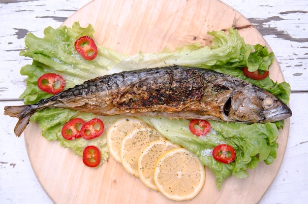 Pesce fritto con fette di limone su insalata di lattuga verde e paprika rossa. Il pesce intero dello sgombro arrostito è servito sul piatto di legno con le verdure