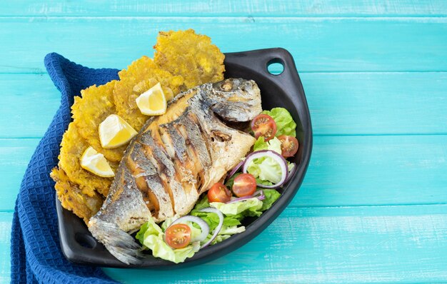 Pesce fritto con insalata e patacones su banda nera su fondo di legno blu. Copia spazio.